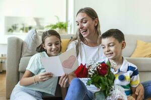 jung Mutter mit ein Strauß von Rosen lacht, umarmen ihr Sohn, und heiter Mädchen mit ein Karte und Rosen gratuliert Mama während Urlaub Feier im Küche beim Zuhause foto