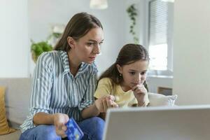 süß klein Tochter Jubel während lächelnd Mutter im beiläufig Kleider Fokussierung auf Bildschirm und mit Laptop zum online Einkaufen im Licht modern Wohnung foto