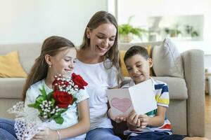 jung Mutter mit ein Strauß von Rosen lacht, umarmen ihr Sohn, und heiter Mädchen mit ein Karte gratuliert Mama während Urlaub Feier im Küche beim Zuhause foto