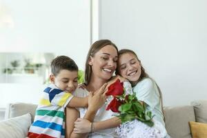 jung Mutter mit ein Strauß von Rosen lacht, umarmen ihr Sohn, und heiter Mädchen mit ein Karte gratuliert Mama während Urlaub Feier im Küche beim Zuhause foto