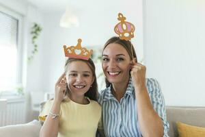 komisch Familie auf ein Hintergrund von hell Mauer. Mutter und ihr Tochter Mädchen mit ein Papier Zubehör. Mama und Kind sind halten Papier Krone auf Stock. foto