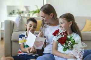 jung Mutter mit ein Strauß von Rosen lacht, umarmen ihr Sohn, und heiter Mädchen mit ein Karte und Rosen gratuliert Mama während Urlaub Feier beim heim. Mütter Tag foto