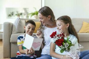 jung Mutter mit ein Strauß von Rosen lacht, umarmen ihr Sohn, und heiter Mädchen mit ein Karte gratuliert Mama während Urlaub Feier im Küche beim Zuhause foto