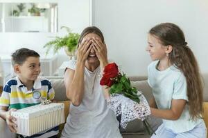 heiter wenig Mädchen mit Strauß von Rosen Blumen und jüngste Bruder mit Geschenk Box lächelnd und Glückwunsch glücklich Mama auf Mutter Tag beim heim. glücklich Mütter Tag foto