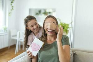 glücklich wenig Mädchen Glückwunsch lächelnd Mutter und geben Karte mit rot Herz während Urlaub Feier beim heim, Abdeckung ihr Augen und überraschend ihr foto