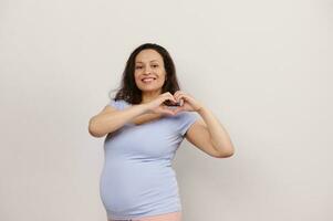 schön lächelnd schwanger Frau Bildung ein Herz gestalten mit Finger, genießen ihr Schwangerschaft und Mutterschaft Lebensstil foto