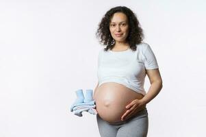 schön Latina schwanger Frau lächelnd beim Kamera, berühren ihr groß Bauch, halten Baby Kleidung, isoliert Weiß Hintergrund foto