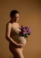 gravid Frau mit schön schwanger Bauch, im Beige Unterwäsche, halten Flieder, erleben glücklich Momente von Schwangerschaft foto