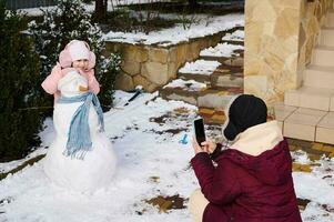 liebend Mutter Fotografien auf Handy, Mobiltelefon Telefon ihr wenig Tochter lächelnd posieren mit Schneemann mit Kopftuch und Karotte Nase foto