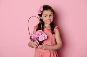 elegant wenig Kind Mädchen tragen Frisur mit Rosen und elegant Rosa Kleid, posieren mit Korb von schön Blumen foto