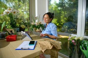 charmant weiblich Florist haben online Video Konferenz mit Klient, Sitzung beim Schreibtisch mit Laptop im Blumen- Design Studio foto