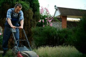 spanisch Gärtner im kariert Blau Hemd und Arbeit Gartenarbeit Uniform, mäht Rasen mit elektrisch Rasen Mäher im Hinterhof foto