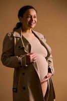 attraktiv gravid Frau, erwartungsvoll Mutter im Beige Graben Mantel und Unterwäsche, lächelnd und sanft streicheln ihr nackt Bauch foto