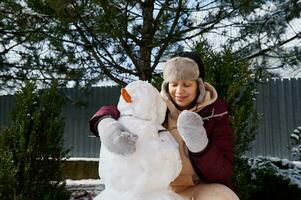glücklich heiter Frau Herstellung ein Schneemann im das Schnee bedeckt Hinterhof, Putten ein Karotte und Baum Ast auf ein enorm Schneeball foto