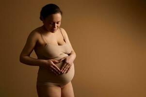 attraktiv jung gravid Frau im Unterwäsche, Bildung Herz geformt Hände auf ihr schwanger Bauch, isoliert Beige Hintergrund foto
