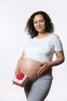 attraktiv positiv multi ethnisch schwanger Frau Mutter mit ein Geschenk Kasten, berühren ihr Bauch, lächelnd suchen beim Kamera foto
