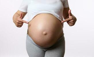 Nahansicht schwanger Frau zeigen Zeigefinger beim ihr groß Bauch im spät Schwangerschaft, isoliert auf Weiß Studio Hintergrund. foto