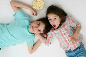 glücklich Kinder mit Popcorn Schüssel Lächeln beim Kamera, ausdrücken Überraschung und Staunen, Lügen Rücken auf Weiß Studio Hintergrund foto