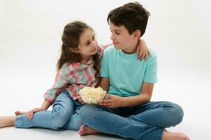 bezaubernd nett Kind Mädchen sanft Umarmungen ihr Bruder Portion ihr ein Schüssel von Popcorn während Film Screening, isoliert auf Weiß foto