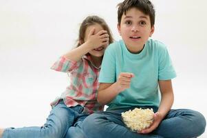 erstaunt Kinder, Teen Junge und wenig Kind Mädchen Aufpassen unheimlich Film oder Karikatur, Essen Popcorn auf Weiß isoliert Hintergrund foto