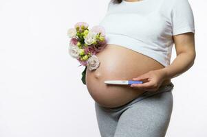 Nahansicht gravid Frau halten ein positiv Schwangerschaft Tintenstrahl Prüfung und Bündel von Frühling Blumen über ihr schwanger Bauch foto