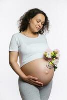 Schwangerschaft. Mutterschaft. Mutter Tag Konzept. charmant schwanger Frau mit Bündel von Blumen, halten Hand auf nackt Bauch foto