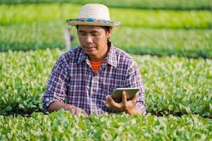 Asiatischer Bauer mit Hut, der ein digitales Tablet verwendet und junge Sämlinge in seiner Farm im Gemüsegarten überprüft