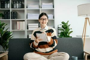 asiatisch Frau mit Laptop und Tablette während Sitzung beim das Sofa. konzentriert beim arbeiten. foto