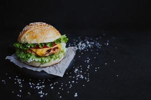 appetitlich frischer hausgemachter Burger, mit Hähnchenschnitzel, Salat, Tomaten, Käse und Sauce. auf einem Holzbrett auf schwarzem Hintergrund foto