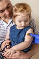 Impfung eines kleinen Mädchens in den Armen ihres Vaters in der Arztpraxis gegen das Coronavirus. lustiges klebepflaster für kinder. Impfung gegen Covid-19, Grippe, Infektionskrankheiten.