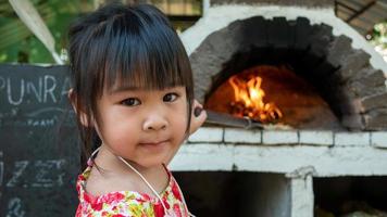 Ein kleines Mädchen macht handgemachte Pizza und steht lächelnd vor einem Pizzaofen aus Holzziegeln in einem Innenhof im Freien in einem Restaurant. foto