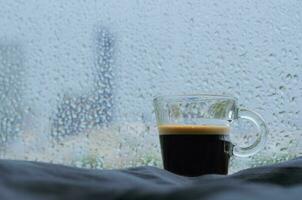 ein Glas von Espresso Kaffee Schuss setzt auf Bett mit Regen fallen auf Fenster im regnet Tag. bleibe Zuhause Konzept. foto