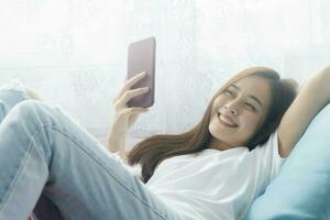 attraktiv lächelnd asiatisch Frau mit Clever Telefon auf das entspannend Sofa beim heim. foto