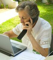 ein alt Frau ist Arbeiten beim ein tragbar Computer und Sprechen auf Handy, Mobiltelefon Telefon foto