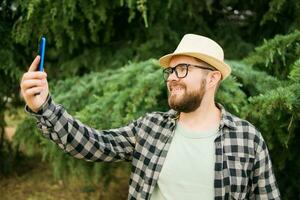 Mann nehmen Selfie Porträt Über Baum Hintergrund - - glücklich tausendjährig Kerl genießen Sommer- Ferien im Stadt - - Jugend und Technologien foto