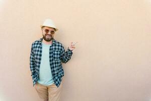 bärtig Mann im Sommer- Kleider und Hut zeigen auf Beige Mauer auf Straße Stadt städtisch und Reise Konzept - - Kopieren Raum und leeren Raum zum Werbung foto