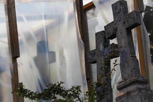 alter Friedhof im Wiederaufbau mit blauem Kreuz hinter einem Busch und mit Plastikfolie auf Holzträgern bedeckt