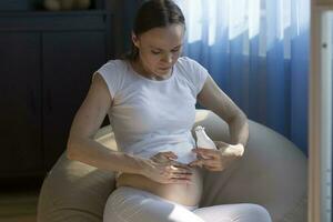 jung schwanger Frau setzt Sahne auf das Bauch Haut foto