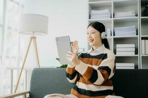 attraktiv glücklich jung asiatisch Schüler studieren beim das Hochschule Bibliothek, Sitzung beim Sofa, mit ein Laptop Computer, Tablette und Kopfhörer haben ein Video Plaudern. foto