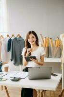 asiatisch Schneider Frau Arbeiten auf Kleider im Schneiderei Atelier. schön jung weiblich Mode Designer Lächeln und nach Erfolg im modern Studio foto