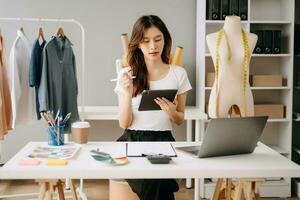 asiatisch Schneider Frau Arbeiten auf Kleider im Schneiderei Atelier. schön jung weiblich Mode Designer Lächeln und nach Erfolg im modern Studio foto