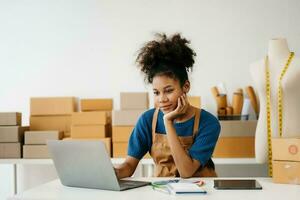 Anfang klein Geschäft klein, Unternehmer Inhaber afrikanisch Frau mit Smartphone oder Tablette nehmen erhalten und Überprüfung online Kauf Einkaufen bestellen zu vorbereiten Pack Produkt Kasten. im Büro foto