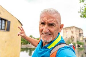 gut aussehend Mitte alt Mann mit Weiß Spitzbart nehmen ein Selfie im das Straßen von ein Italienisch Dorf durch das See foto