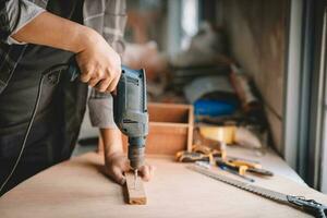 schließen oben von männlich Mechaniker halten elektrisch schnurlos Schraubendreher bohren mit Holz Schraube im das Fabrik. Arbeiten mit das schrauben. Fachmann Tischler. foto