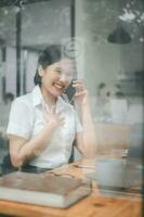 attraktiv jung Frau reden auf das Handy, Mobiltelefon Telefon und lächelnd während Sitzung beim ihr Arbeiten Platz im Büro und suchen beim Laptop foto
