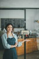 Porträt von glücklich Kellnerin Stehen beim Restaurant Eingang mit öffnen Zeichen, Porträt von jung Geschäft Frau besuchen Neu Kunden im ihr Kaffee Geschäft. foto