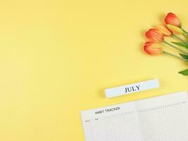 eben Layout von Gewohnheit Tracker Buch, hölzern Kalender Juli und Tulpen auf Gelb Hintergrund mit Kopieren Raum. foto