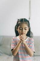 wenig asiatisch Mädchen beten mit ihr Hände umklammert zusammen beim Zuhause foto
