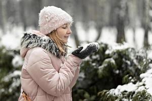 glückliche Frau an einem verschneiten Wintertag im Park, in warme Kleidung gekleidet, bläst den Schnee von ihren Handschuhen