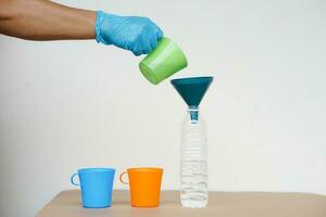 Nahansicht Hand tragen Blau Handschuh hält Grün Becher zu gießen Wasser in Flasche zu tun Wissenschaft Experiment. Konzept, Wasser Menge Messung zum Mischen chemisch Substanzen. foto